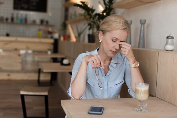 Tired middle aged women pinching the bridge of her nose sitting at a cafe with a drink and phone on table